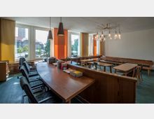 Ein heller Raum mit großen Fenstern, die Tageslicht hereinlassen, ausgestattet mit langen Holztischen, Stühlen und einer Regalwand mit Büchern. Im Vordergrund steht ein geschwungener Holztisch mit Büromaterialien, und an der Decke hängen moderne Leuchten.