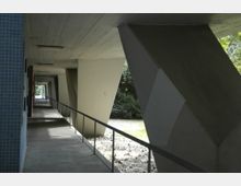 Überdachter Gehweg mit modernem Betonpfeiler-Design, flankiert von einem Geländer auf der linken Seite und einer Reihe massiver schräger Stützen rechts; im Hintergrund Bäume und eine Wiese.