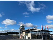 Flughafen mit Kontrollturm und angrenzendem Gebäude mit Glasfassade und Terrasse, daneben wehen mehrere Flaggen. Im Hintergrund sind ein Hangar, kleine Flugzeuge und ein bewaldeter Horizont zu sehen, unter einem blauen Himmel mit Wolken.
