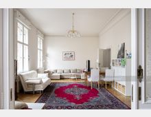 Wohnzimmer in einem Altbau mit hohen Decken, großen weißen Fenstern und einem hölzernen Parkettboden. Der Raum ist möbliert mit einer beigen Eckcouch, einem Sessel, einem roten Orientteppich sowie einem Esstisch mit weißen Stühlen; ein Regal steht an der Wand.