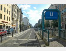 Straßenszene an der U-Bahn-Station Oranienburger Tor in Berlin mit gelbem Tramgleis, Gehwegen, mehrstöckigen Häusern, Geschäften und einer Bushaltestelle. Rechts im Bild führt eine Treppe mit Glasgeländer zur U-Bahn, markiert durch ein blaues Schild mit dem Buchstaben "U".