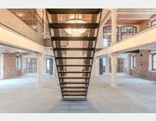 Industrie-Loft mit sichtbaren Backsteinwänden, weißen Pfeilern, großen Metall-Fenstern und einer modernen schwarzen Metalltreppe. Über der Treppe hängt ein großer Kronleuchter in einem offenen, hellen Raum mit hohen Decken.