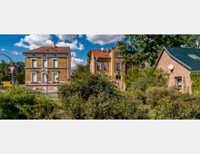 Straßenszene mit historischen zweigeschossigen Gebäuden aus gelben und roten Ziegeln sowie einem modernen eingeschossigen Haus mit grünem Dach im Vordergrund. Im unteren Bildbereich sind Büsche und Büten zu sehen, im Hintergrund Bäume unter klarem Himmel mit weißen Wolken.