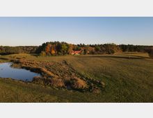Weite Landschaft mit einer kleinen Wasserfläche im Vordergrund, umgeben von Gräsern und Wiesen, im Hintergrund ein einzelnes Gebäude mit rotem Dach, umgeben von herbstlich gefärbten Bäumen und Wald.