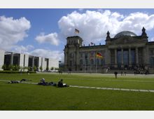 Das Bild zeigt das Reichstagsgebäude in Berlin mit seiner markanten Glaskuppel. Im Vordergrund liegt eine große Rasenfläche mit Menschen, die entspannen, während im Hintergrund moderne Bürogebäude und wehende Flaggen zu sehen sind.