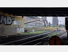 Blick auf Bahngleise unter einer Brücke mit Graffiti an der Wand und moderner Architektur im Hintergrund, darunter Gebäude mit Glas- und Stahlfassaden sowie Hochhäuser. Im Hintergrund sind Kräne und Baustellen sichtbar.