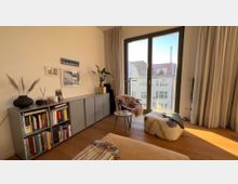 Gemütliches Wohnzimmer mit Holzfußboden, einem grauen Regal voller Bücher und Dekorationen, einem Stuhl mit Fellüberwurf und einem kleinen Tisch. Neben einer großen Fensterfront mit Blick auf Nachbarhäuser und einen Turm fällt Tageslicht in den Raum, flankiert von beigen Vorhängen.