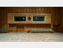 Großer Raum mit hölzerner Wandverkleidung, zwei schallisolierten Fenstern, einer zentralen analogen Uhr und einer großen hölzernen Tür darunter. Der Boden ist mit Fischgrätparkett ausgelegt, und entlang der Wände stehen grüne Polstersofas.