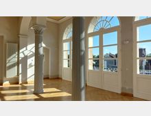 Heller Innenraum mit hohen Fenstern und Rundbögen, teilweise durch elegante Säulen unterteilt. Der Raum hat helle Wände, einen Parkettboden und lässt durch die Fenster den Blick auf ein Gebäude und Bäume im Außenbereich zu.