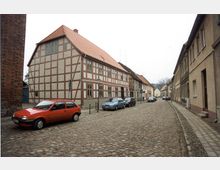 Straßenszene in einer historischen Altstadt mit Kopfsteinpflaster, Fachwerkhaus mit roten Dachziegeln auf der linken Seite und beige verputzten Gebäuden rechts; mehrere parkende Autos säumen die Straße.