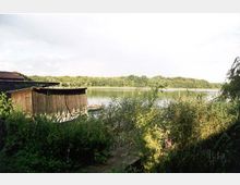 Holzhütte am Ufer eines ruhig wirkenden Sees, umgeben von dichtem Schilf und Vegetation; im Hintergrund sind Wälder am gegenüberliegenden Ufer zu sehen, unter einem hellen, teilweise bewölkten Himmel.