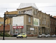 Straßenansicht eines historischen Gebäudes mit grauer Fassade und abgenutztem Putz in Berlin-Köpenick, mit mehreren Werbetafeln, darunter "Zum Waschhaus Alt-Köpenick". Vor dem Gebäude parken Autos, und ein Straßenschild mit der Aufschrift „Alt-Köpenick“ ist im Vordergrund sichtbar.