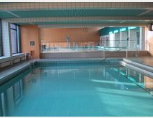 Hallenbad mit einem rechteckigen Becken im Vordergrund, umgeben von gefliesten Wänden und Böden. Im Hintergrund befindet sich ein weiteres höher gelegenes Becken, getrennt durch eine Glaswand, sowie Umkleidebereiche und große Fenster, die natürliches Licht hereinlassen.