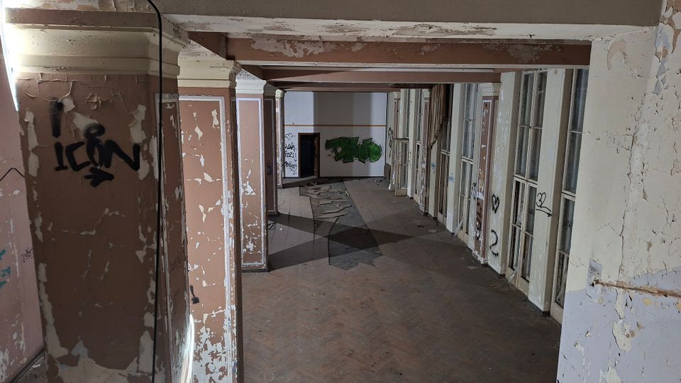 Verlassener Innenflur eines alten Gebäudes mit abblätternder Farbe, Stützpfeilern und freiliegenden Deckenbalken. Rechts verläuft eine Reihe hoher Fenster- oder Türrahmen, an Wänden und Türen sind Graffiti und Risse im Putz zu sehen.