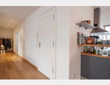 Flur mit Holzfußboden und weiß gestrichenen Wänden, links eine Garderobe mit Kleidungsstücken, rechts eine offene Küche mit Regal, Kupferkannen, Kochbüchern und einer Dunstabzugshaube. Eine weiße Tür mit goldfarbener Klinke befindet sich in der Mitte.