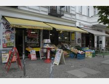Straßenansicht eines kleinen Ladens mit gelben Markisen, auf dem „Tabak“, „Presse“ und „Getränke“ steht. Vor dem Geschäft stehen Verkaufsständer mit Obst und Gemüse sowie Werbetafeln, daneben ein weiterer Eingangsbereich mit ähnlichem Warenangebot.