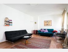 Wohnzimmer mit Holzboden, einem schwarzen Sofa, einem türkisfarbenen Sofa mit lila Kissen, einem kleinen Tisch und einem bunten Teppich. Eine Wand ist mit schwebenden Bücherregalen und abstrakter Kunst dekoriert, helle Vorhänge rahmen ein Fenster rechts im Raum.