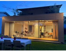 Moderne Wohnhausfassade mit großen Glasfenstern, hinter denen ein beleuchtetes Wohnzimmer und eine Küche sichtbar sind. Im Vordergrund befindet sich ein Garten mit einem Esstisch und mehreren Stühlen auf einer Rasenfläche.