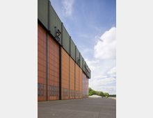 Großes Hangargebäude mit orangefarbenen Metallpaneelen und grüner Dachverkleidung, umgeben von freiem Asphalt- und Grünflächen unter einem teils bewölkten blauen Himmel.
