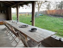 Eine überdachte Terrasse mit einem langen rustikalen Holztisch und mehreren Holzklappstühlen, die auf einen grünen Garten mit Bäumen und einem Zaun im Hintergrund blickt. Die Decke und Pfosten bestehen aus Holz, und der Raum ist teils offen mit Blick auf die ländliche Szenerie.