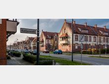 Straßenszene mit historischen Backsteinhäusern, roten Dächern und kleinen Balkonvorsprüngen, die entlang einer Straße mit geparkten Autos stehen. Im Vordergrund Straßenschilder mit den Namen "Kastanienstraße" und "Karl-Marx-Straße".