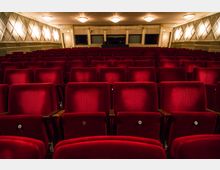 Innenansicht eines Theatersaals mit mehreren Reihen gepolsterter, roter Samtsitze. Der Raum hat dezente Wandleuchten, ein helles Deckenmuster und Kronleuchter, die den Saal gleichmäßig ausleuchten.