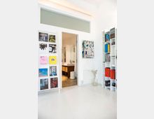 Minimalistisch gestalteter Innenraum mit weißen Wänden und einem glänzend weißen Boden. Links steht ein Regal mit farbigen Magazinen, rechts ein Bücherregal mit farblich geordneten Büchern, in der Mitte eine Tür, die zu einem Badezimmer mit Waschbecken und Spiegel führt.