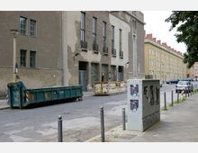 Straßenansicht einer grauen, verputzten Gebäudefassade mit hohen, schmalen Fenstern und Gittern im Erdgeschoss. Im Vordergrund stehen Container, ein Stromkasten mit Plakaten sowie Poller, und die Umgebung zeigt parkende Autos und eine weitere Häuserzeile im Hintergrund.
