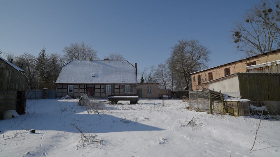 Verschneiter Hof eines ländlichen Anwesens mit einem Fachwerkhaus und schneebedecktem Satteldach in der Mitte, umgeben von Nebengebäuden und Zäunen. Im Vordergrund liegt eine offene, schneebedeckte Fläche mit verstreuten Pflanzenresten und Spuren, dahinter stehen kahle Bäume vor klarem Himmel.