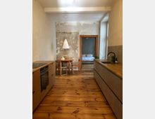 Küche in minimalistischem Stil mit Holzfußboden, hellen Holzschränken und einer Arbeitsplatte. Im Hintergrund ein Essbereich mit rundem Holztisch und Stühlen vor einer Wand mit sichtbaren Putzspuren, durch die offene Tür ein Schlafzimmer erkennbar.