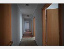 Langer, schmaler Innenflur mit weißen Wänden und mehreren Holztüren auf beiden Seiten. An der Decke hängen einfache Leuchten, der Boden wirkt teils unfertig und am Ende des Gangs ist eine offene Tür mit Hakenleiste sichtbar.