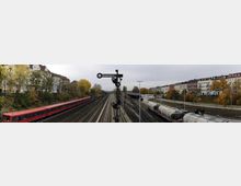 Blick auf ein Bahnhofsgebiet mit mehreren Gleisen, auf denen links ein roter Zug und rechts Güterwagen zu sehen sind. Im Hintergrund befinden sich eine Baumreihe mit herbstlich verfärbten Blättern sowie Wohngebäude entlang der Strecke.