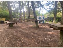 Ein Kletterpark im Wald mit hölzernen Plattformen und Seilelementen, die zwischen Bäumen aufgehängt sind. Im Hintergrund stehen tragbare Toiletten und Sitzbereiche mit Tischen, umgeben von dichtem Grün.