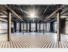 Großer, industrieller Raum mit einem schwarz-weißen Schachbrettmusterboden, Metallträgern an der Decke und hohen Fenstern mit Sprossen, die viel Tageslicht hineinlassen. Die Wände bestehen aus hellen Backsteinen, und an den Seiten befinden sich schwarze Vorhänge.