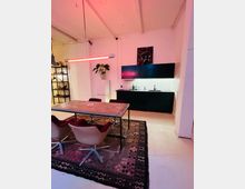 Wohnbereich in einem modernen Industriestil, mit einem Holztisch und vier gepolsterten Stühlen auf einem gemusterten Teppich, einer schwarzen Küchenzeile mit weiß gefliester Rückwand sowie einer roten Hängelampe. Im Hintergrund steht ein Regal und eine Zimmerpflanze.