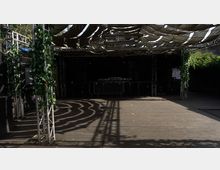 Holzboden-Terrasse mit einer Bühne im Hintergrund und einem Überdachungssystem aus Stoffbahnen. Metallkonstruktionen mit grünem Blattdekor säumen die Seiten, umgeben von einigen Pflanzen.