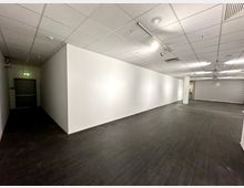 Schlichter, leerer Innenraum mit weißen Wänden, dunklem Boden und abgehängter Decke mit integrierter Beleuchtung. Im Hintergrund sind ein geschlossenes Rolltor und eine Notausgangstür sichtbar.