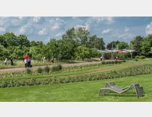 Ein gepflegter Park mit Blumenbeeten, Rasenflächen und Spazierwegen, umgeben von Bäumen. Im Hintergrund sieht man eine kleine Überdachung mit Sitzgelegenheiten, während Personen entlang des Weges spazieren.