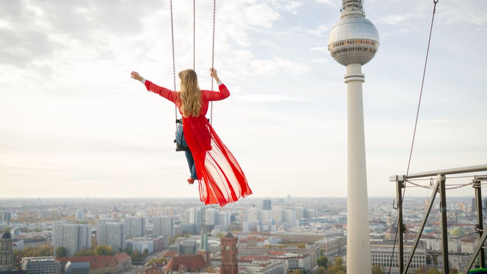 Blick von einer hohen Plattform über die Berliner Innenstadt mit dem Fernsehturm rechts im Bild. Im Vordergrund sind Stahlstreben, Seile und eine hängende Schaukel über dem Stadthorizont zu sehen.