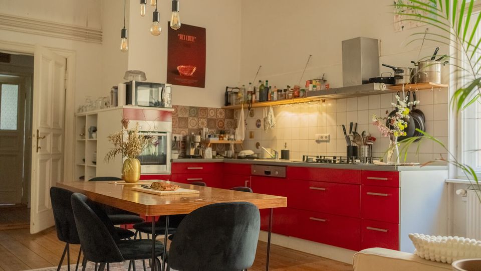 Open-plan kitchen and dining area with red lower cabinets, white tiled backsplash, and a long countertop with stove and range hood. A wooden dining table with black chairs sits under hanging pendant bulbs, with open shelving, small appliances, and potted greenery near a large window.