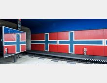 U-Bahn-Station mit einer Wand, die mit einem Muster gestaltet ist, das der norwegischen Flagge ähnelt. Die Szene zeigt das Gleis mit einer Signaltafel und die gepflasterte Plattform im Vordergrund.