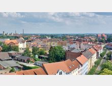 Stadtansicht mit roten Ziegeldächern, mehrstöckigen Wohnhäusern, einer Kirche am Horizont und Fabrikschornsteinen; im Hintergrund eine flache Landschaft unter leicht bewölktem Himmel.
