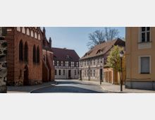 Straßenszene in einer Altstadt mit historischen Gebäuden aus Backstein und Fachwerk, gepflasterter Straße und Laternen. Im Hintergrund sind kahle Baumkronen und ein klarer Himmel zu sehen.