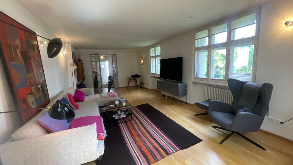 Helles Wohnzimmer mit Holzfußboden, großem Sofa mit Couchtisch und farbigem Teppich sowie einem Fernseher auf einem Lowboard. Rechts sorgen hohe, mehrteilige Fenster für viel Tageslicht; daneben steht ein gepolsterter Sessel, im Hintergrund führt eine Glastür in den nächsten Raum.