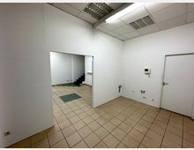 Leerer Raum mit weißen Wänden, beige-braunen Fliesenboden und abgehängter Decke mit Rasterleuchten. Im Hintergrund ein Durchgang zu einem weiteren Raum mit einer kleinen Treppe, und rechts eine weiße Tür.