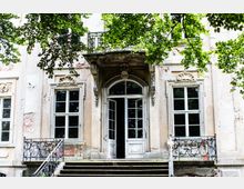 Historische Fassade eines Gebäudes mit verzierten Fenstern und einem großen Eingangsportal, umgeben von schmiedeeisernen Geländern. Im Vordergrund sind Treppenstufen und überhängende Äste von Bäumen sichtbar.