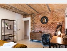 Schlafzimmer mit rustikaler Einrichtung, sichtbarer Ziegelwand und Gewölbedecke. Es gibt ein hölzernes Sideboard, einen Spiegel, ein Regal mit schwarzem Metallrahmen, einen Schreibtisch mit Tischlampe und einen Stuhl mit Fellüberzug.