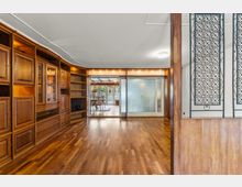 Großer Wohnraum mit Parkettboden und einer wandfüllenden, eingebauten Holzschrankwand mit Regalen und Vitrinentüren. Im Hintergrund führen breite Schiebetüren aus Glas in einen angrenzenden Essbereich; rechts ist eine erhöhte Nische mit Holzvertäfelung und dekorativem Metallgitter zu sehen.