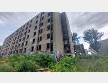 Ein verlassenes, mehrstöckiges Wohngebäude mit zerstörten Fenstern und Graffiti an den Wänden. Umgeben von dichter Vegetation und vereinzelten Bäumen unter einem bewölkten Himmel.