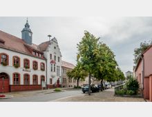 Straßenszene mit einem historischen Rathaus links im Bild, gekennzeichnet durch weiße Fassade, rote Fensterrahmen und einen kleinen Turm mit Uhr. Rechts neben der Straße stehen Bäume und geparkte Autos, während auf der rechten Seite weitere Gebäude und ein kleiner Zaun mit Grünpflanzen zu sehen sind.